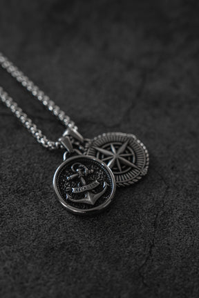 Two silver stainless steel necklaces from Relentless Betrayal: the Reckless Soul Necklace with an anchor engraved RECKON, and a compass pendant. Both handcrafted pieces rest on a dark textured surface.
