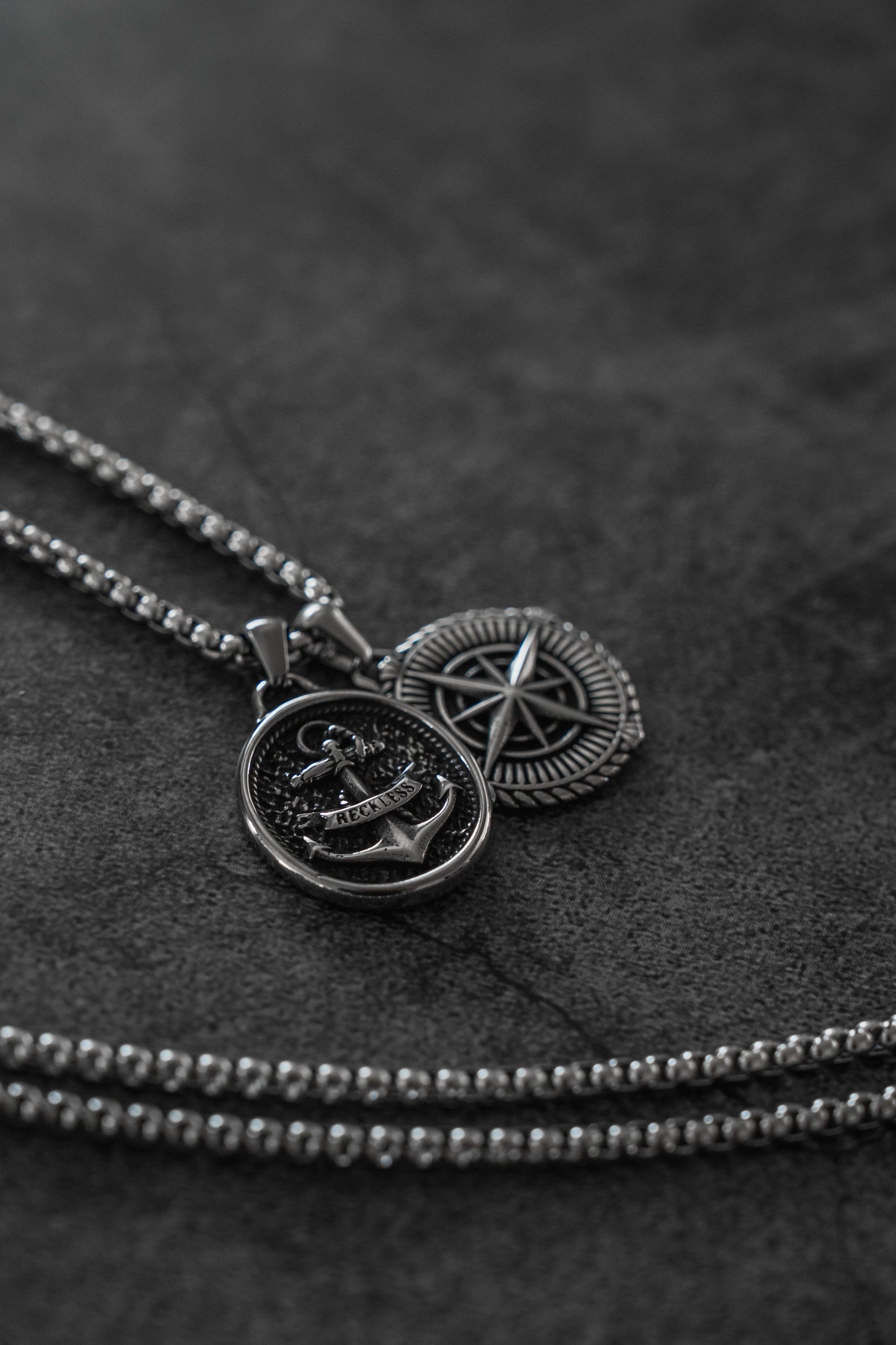 Two Reckless Soul Necklaces by Relentless Betrayal—a silver anchor with rope and a star in a circle—rest on a dark textured surface. Each handcrafted stainless steel piece comes with a 26-inch chain.