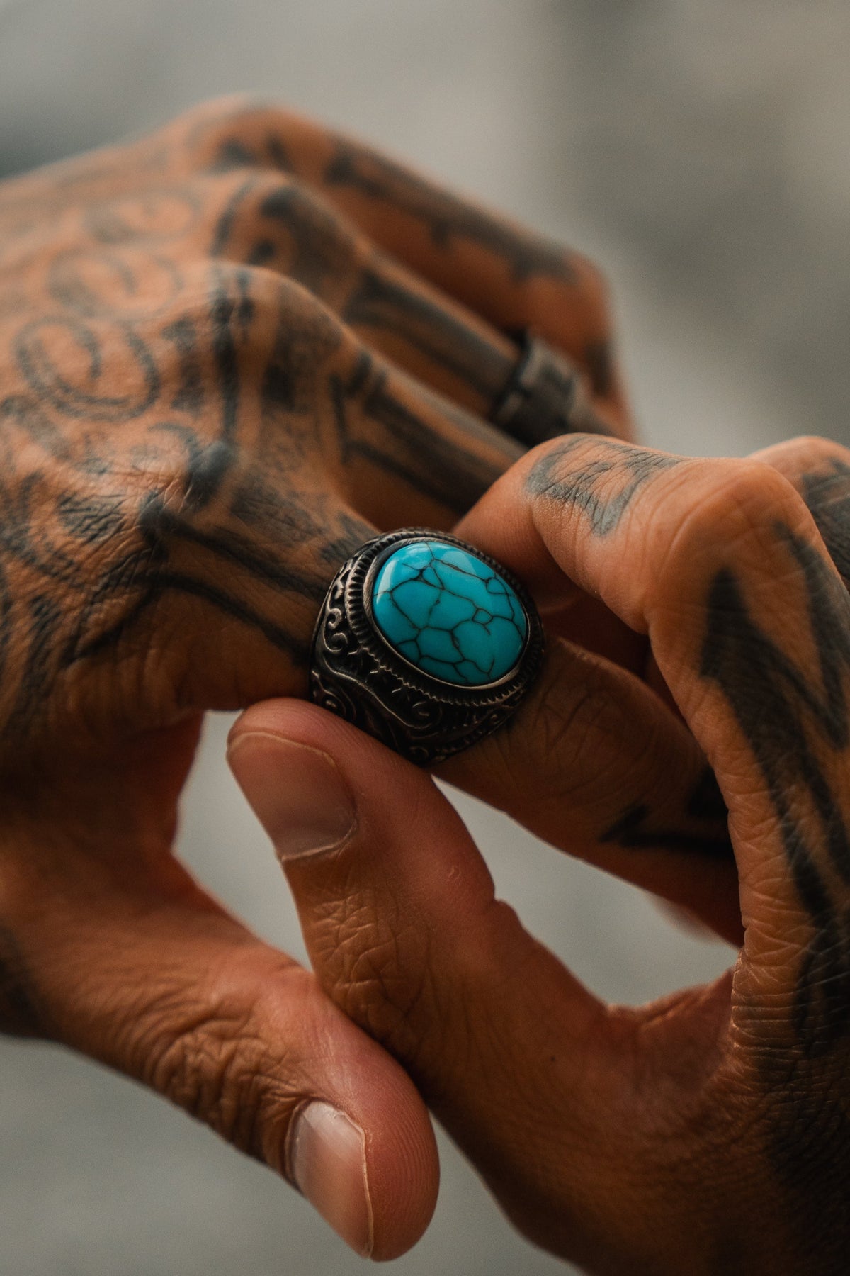 A tattooed hand puts on the handcrafted Into the Storm Ring by Relentless Betrayal, featuring an ornate stainless steel band and a bright turquoise stone, while another tattooed hand assists. The background is blurred.