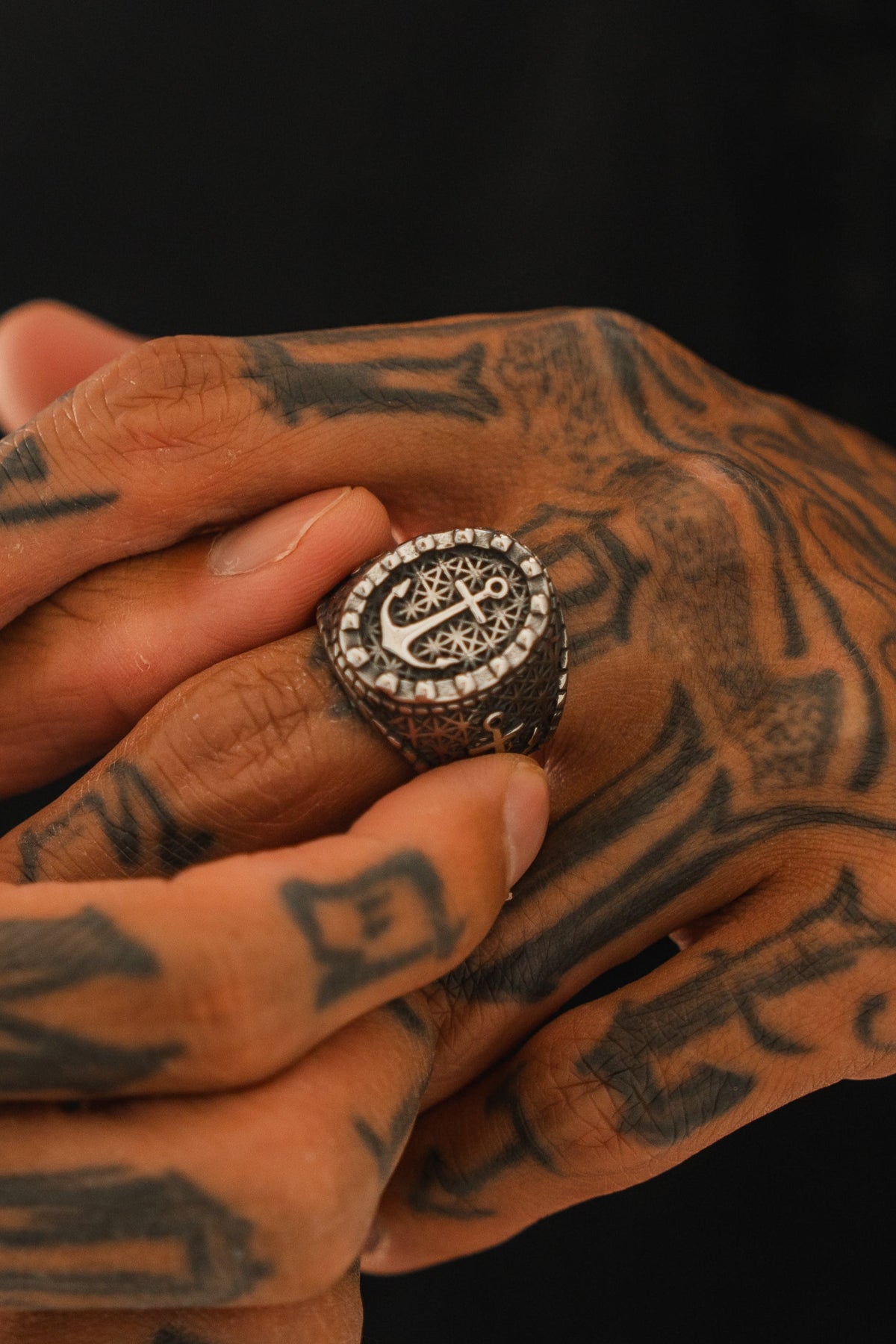 Tattooed hands hold the Anchored in Glory Ring by Relentless Betrayal, featuring intricate designs and patterns. Set against a dark background, it highlights the bold style of nautical jewelry.