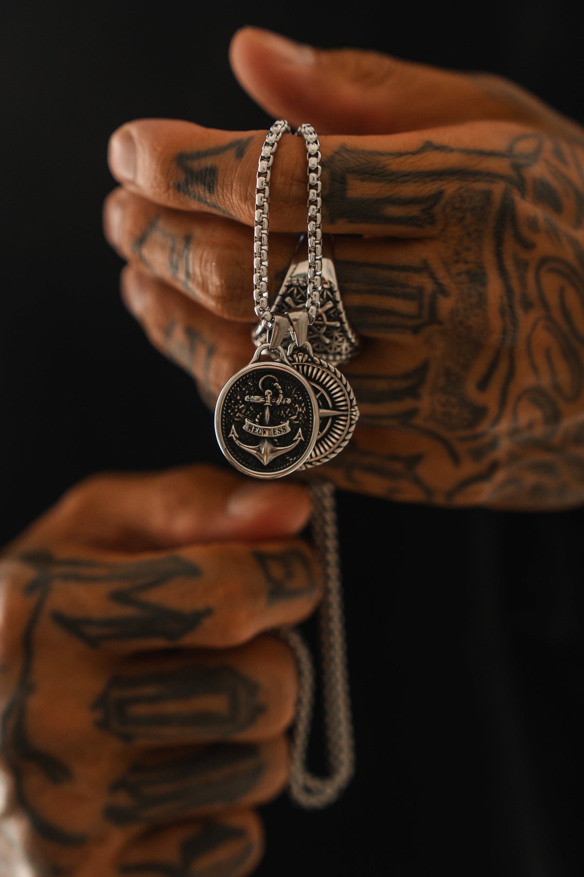 A tattooed hand holds two silver pendant necklaces, including the handcrafted Relentless Betrayal Reckless Soul Necklace featuring an anchor design, against a dark background.