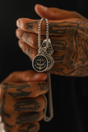A tattooed hand holds two silver pendant necklaces, including the handcrafted Relentless Betrayal Reckless Soul Necklace featuring an anchor design, against a dark background.