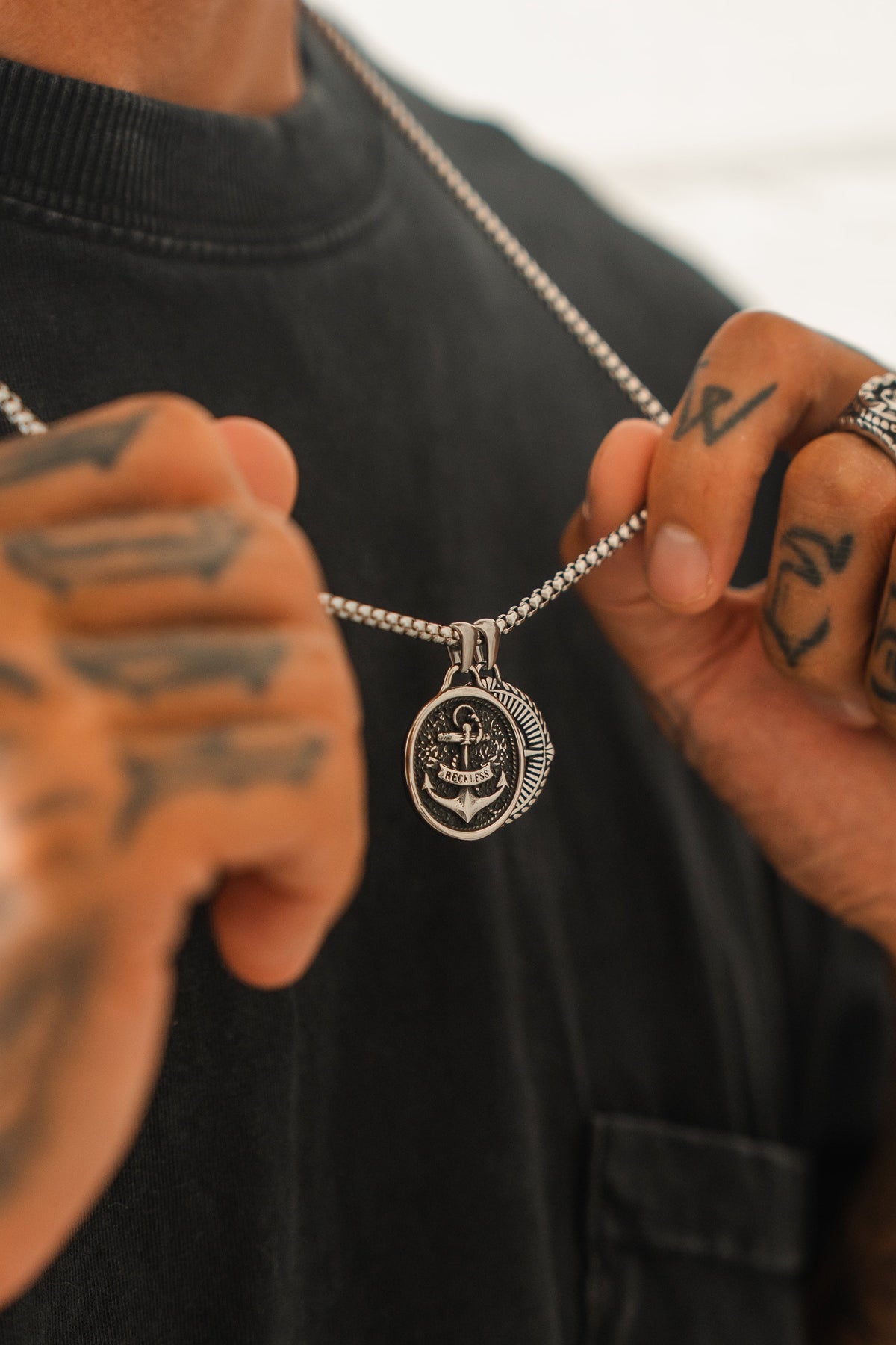 A tattooed hand with rings holds the Relentless Betrayal Reckless Soul Necklace—a handcrafted stainless steel piece with a round anchor-engraved pendant—while wearing a dark shirt.
