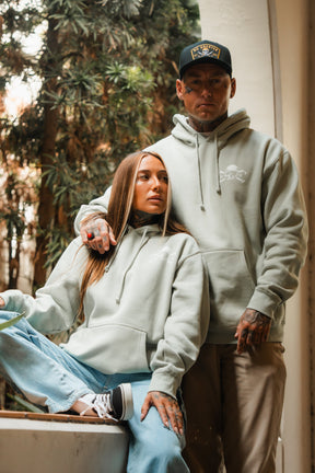 Two people wear Relentless Betrayal’s Tell No Tales Premium Sage Hoodie—one seated in blue jeans and sneakers, the other standing in khaki pants and a black cap—posing outdoors amid greenery.