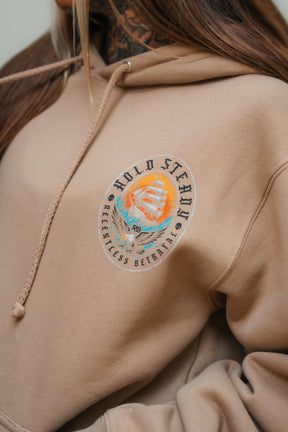 A person wears the Hold Steady Premium Hoodie by Relentless Betrayal, featuring a tan heavyweight fabric and an embroidered sailing ship patch on the chest. Only part of their face and hair is visible, highlighting this unique unisex hoodie.