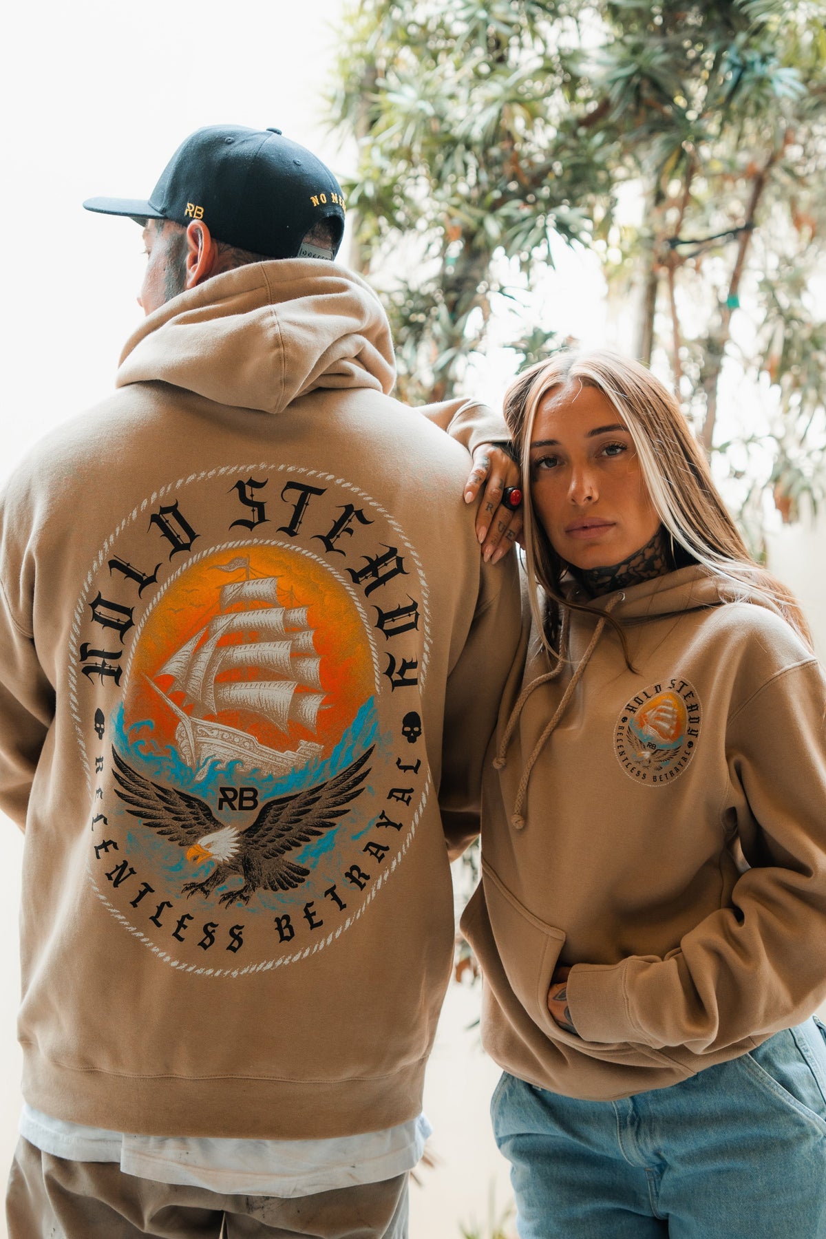 Two people wear Relentless Betrayal Hold Steady Premium Hoodies outdoors. The woman faces forward, while the man shows off the ship graphic and Relentless Betrayal text on the back among leafy trees.
