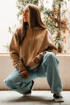 A woman with long brown hair, wearing the Relentless Betrayal Hold Steady Premium Hoodie, light blue baggy jeans, and black-and-white sneakers, kneels outside before leafy plants, gazing to the side with a relaxed expression.