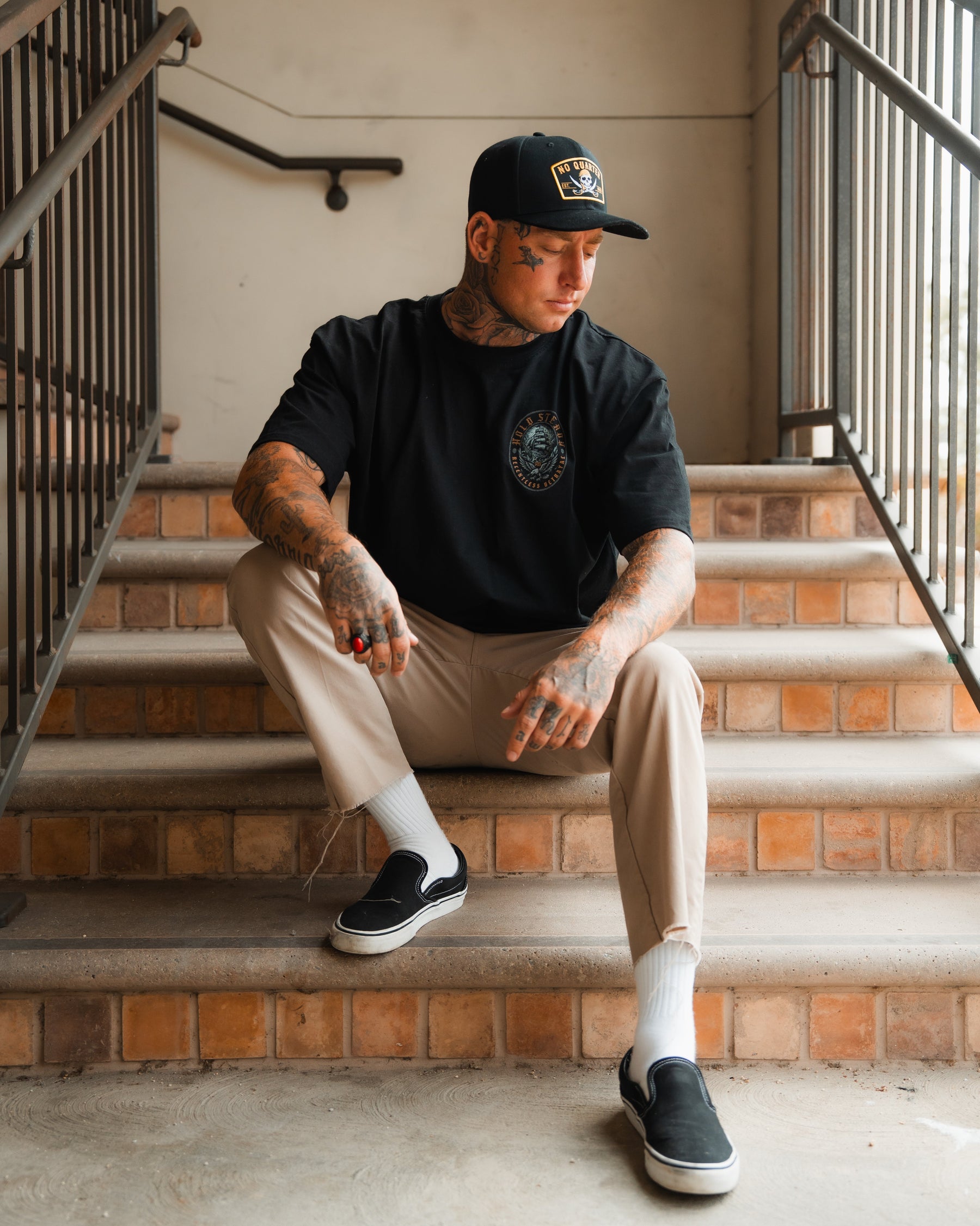 A tattooed man in a black cap and t-shirt, beige pants, and black slip-ons sits on stairs, wearing the Relentless Betrayal Burning Rose Blackout Ring, with one arm on his knee that shows off his tattoos.