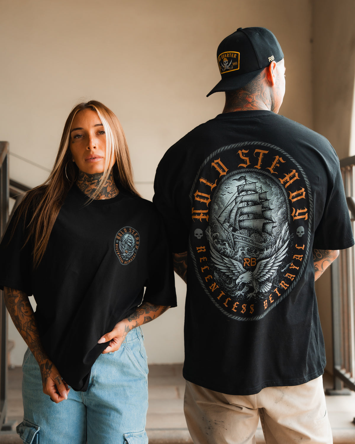 Two tattooed models wear the Relentless Betrayal Hold Steady Premium Oversized Tee—she faces forward in black with a small chest print, while he shows off the bold ship graphic and lettering featured on the back.