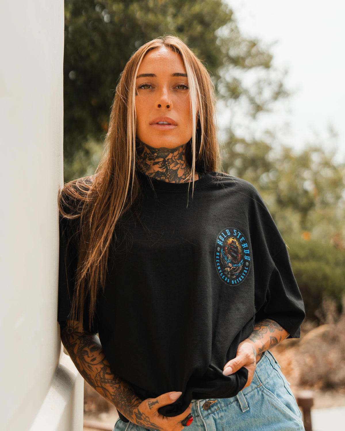 A woman with long brown hair and visible tattoos leans against an outdoor wall, wearing the Relentless Betrayal Hold Steady Blue - Premium Oversized Tee and light blue jeans, with blurred greenery in the background.
