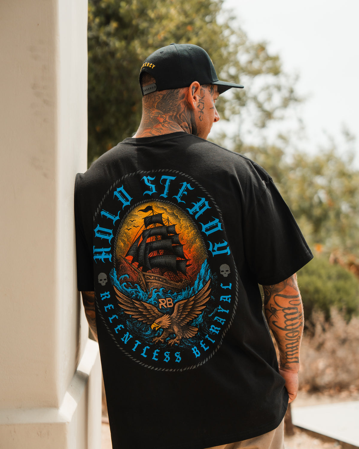 A tattooed person outdoors wears the Relentless Betrayal Hold Steady Blue Premium Oversized Tee, a black shirt featuring a ship and eagle graphic with bold blue and orange text.