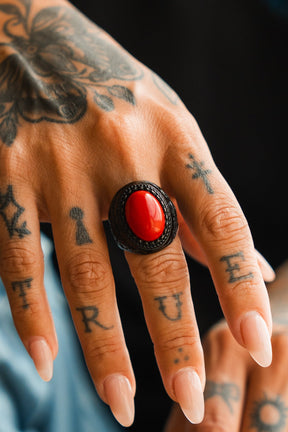 A tattooed hand with "TRUE" inked across the fingers wears the Relentless Betrayal Burning Rose Blackout Ring, showcasing a bold red stone. Long manicured nails and additional tattoos complete the striking look.