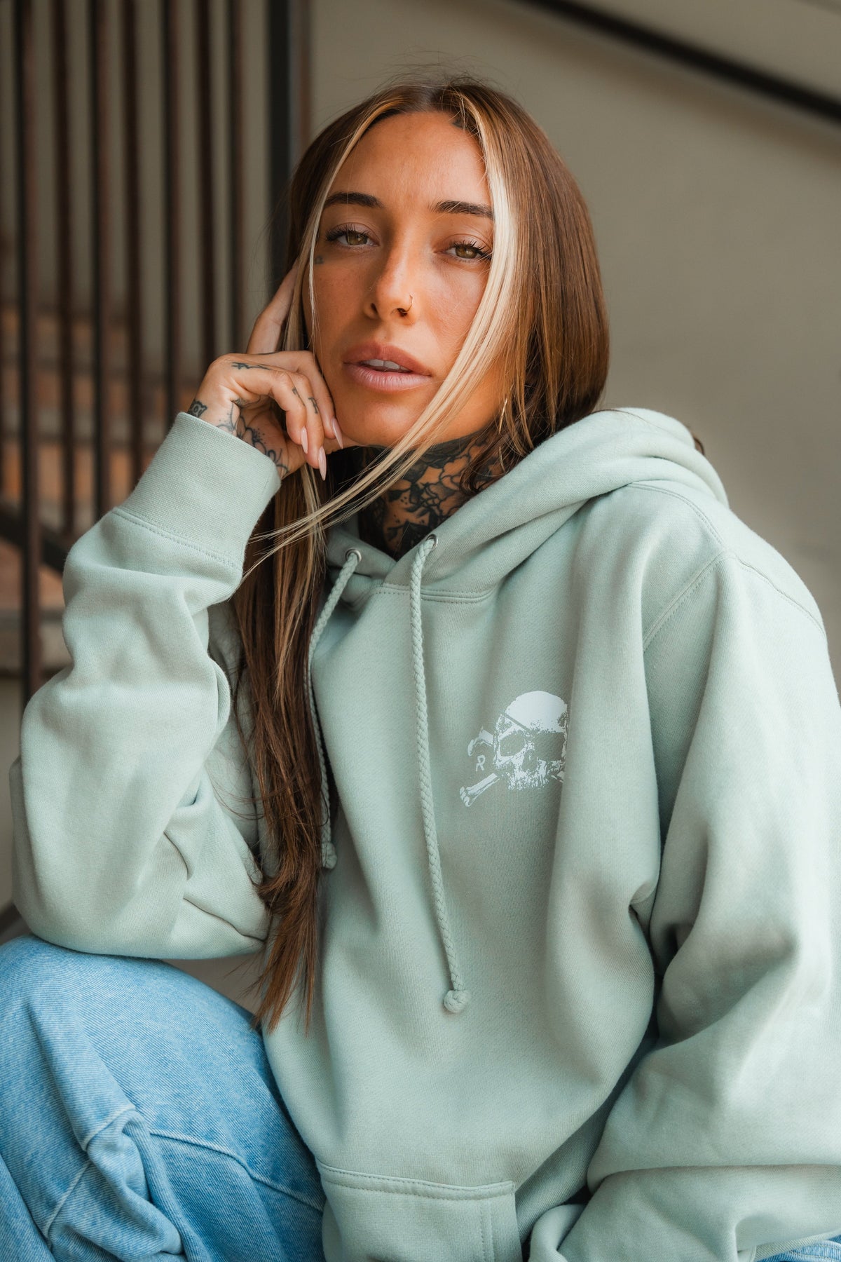 A woman with long, light brown hair and tattoos sits on stairs wearing the Relentless Betrayal Tell No Tales Premium Sage Hoodie with a small skull graphic and light blue jeans, resting her head on her hand and gazing at the camera.