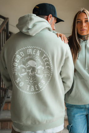 A man in a Relentless Betrayal Tell No Tales Premium Sage Hoodie stands next to a woman in a matching cotton-polyester blend hoodie. Both display tattoos and relaxed expressions as they pose indoors near stairs.