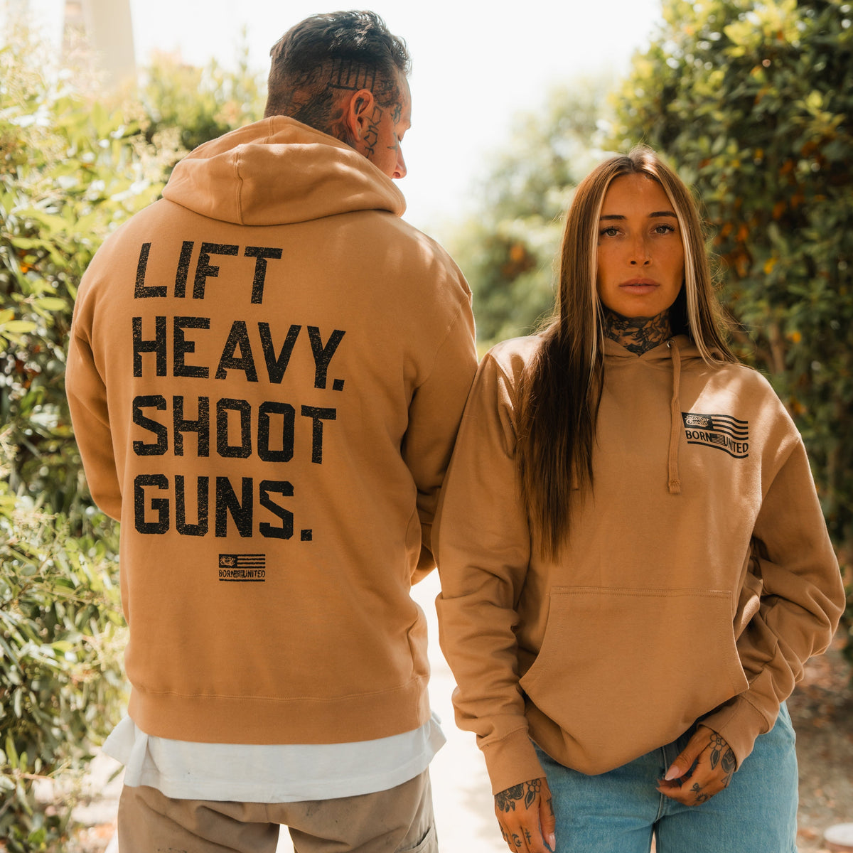 A man and woman in tan hoodies stand outdoors, both with tattoos and neutral expressions. The mans hoodie is the LHSG - Hoodie by Tango Charlie Apparel, featuring the bold Lift Heavy Shoot Guns motto—perfect for fitness and 2A enthusiasts.