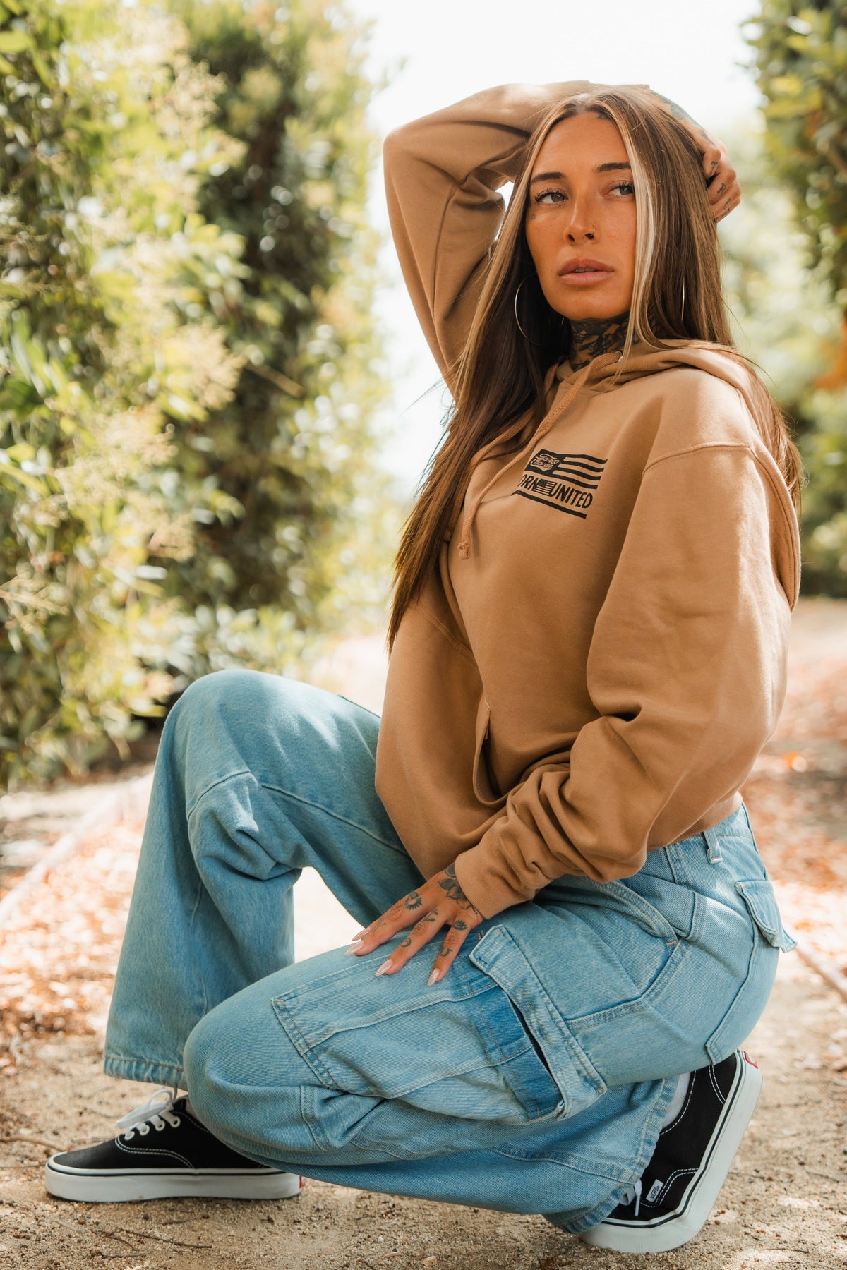 A woman with long hair kneels outdoors on a dirt path, surrounded by greenery. She wears the LHSG - Hoodie from Tango Charlie Apparel, paired with light blue cargo jeans and black sneakers, resting one hand on her head in thought.