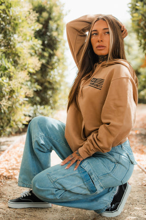 A woman with long hair kneels outdoors on a dirt path, surrounded by greenery. She wears the LHSG - Hoodie from Tango Charlie Apparel, paired with light blue cargo jeans and black sneakers, resting one hand on her head in thought.