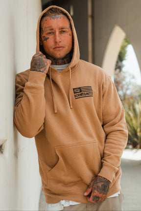 A tattooed man stands outdoors against a wall, wearing the LHSG - Hoodie by Tango Charlie Apparel—a tan high-intensity fitness hoodie with “Born United” text—holding his hood with one hand.