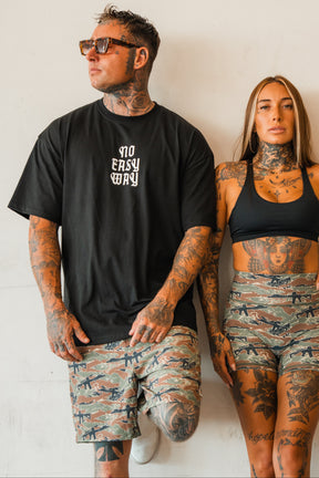 A tattooed man and woman stand together by a plain wall. He wears sunglasses, a black tee, and Tango Charlie Apparel Men's Performance Training Shorts | 5.5"- Snipes & Stripes. She pairs her black crop top with matching camo gym shorts.