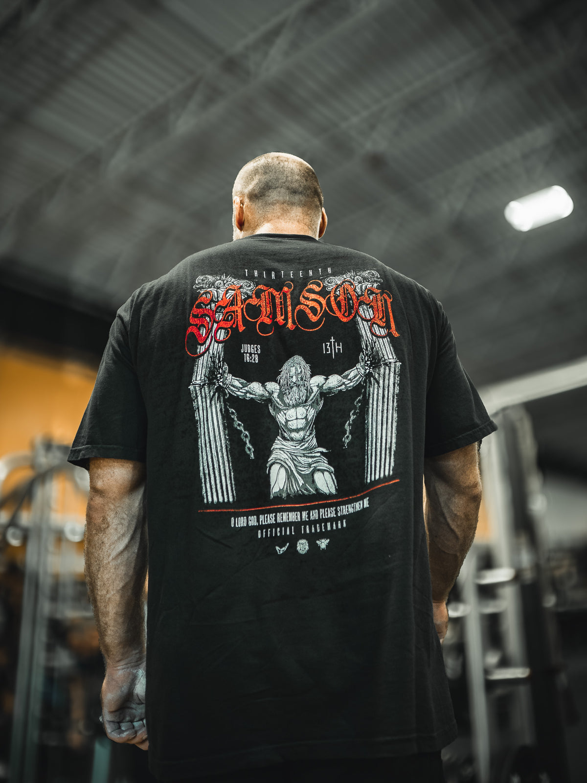 A muscular man in a gym wears the 13th Samson Premium Tee, featuring bold red and white designs of Samson breaking chains, with gym equipment blurred in the background.