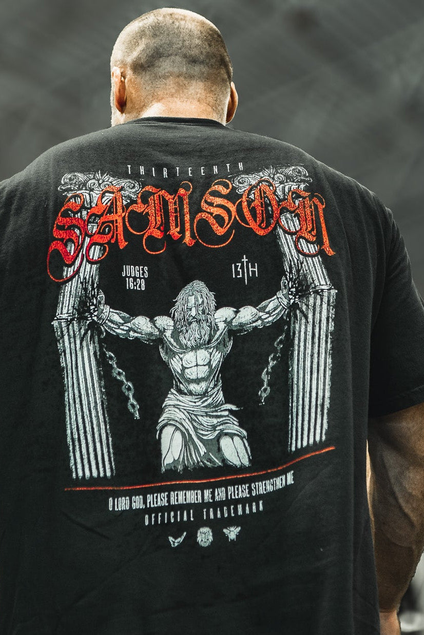 A muscular man in a gym wears the 13th Samson Premium Tee, featuring bold red and white designs of Samson breaking chains, with gym equipment blurred in the background.