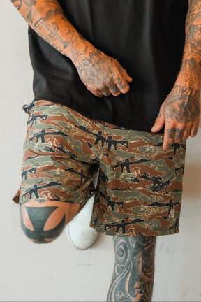 A person with heavily tattooed arms and legs wears Tango Charlie Apparel's Men's Performance Training Shorts | 5.5"- Snipes & Stripes, a black shirt, and white shoes, standing against a light wall with one knee bent.