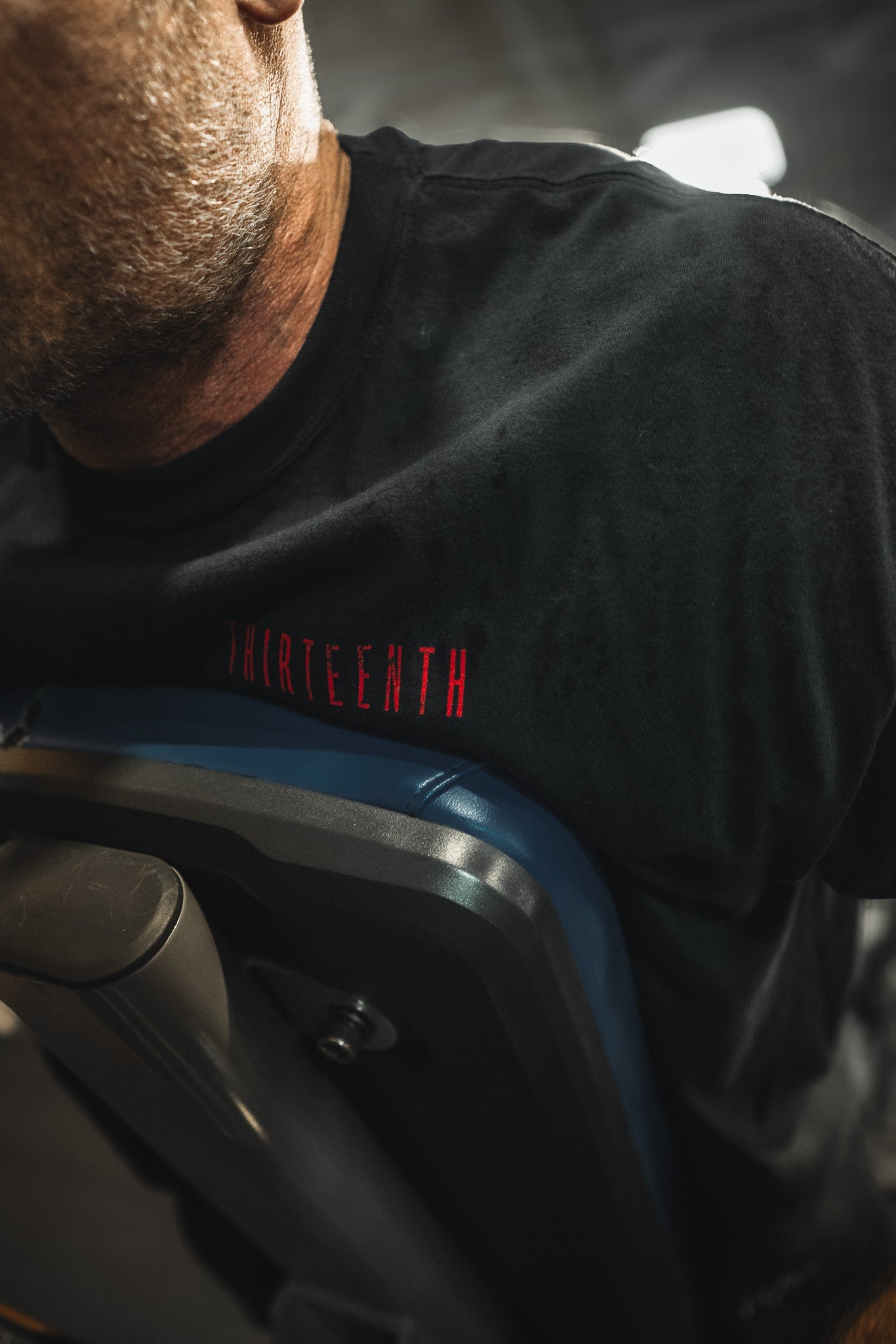 A man wearing the black 13th Samson Premium Tee with THIRTEENTH in red exercises on gym equipment, with the image highlighting his upper torso and part of his face.
