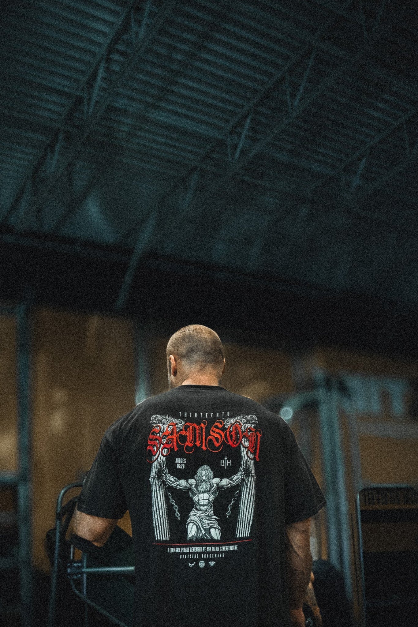 A person with a shaved head stands indoors, facing away, wearing the 13th Samson Premium Tee featuring a bold graphic and red text on the back. The dim lighting and industrial setting accentuate this premium garment.