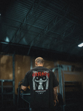 A person with a shaved head stands indoors, facing away, wearing the 13th Samson Premium Tee featuring a bold graphic and red text on the back. The dim lighting and industrial setting accentuate this premium garment.