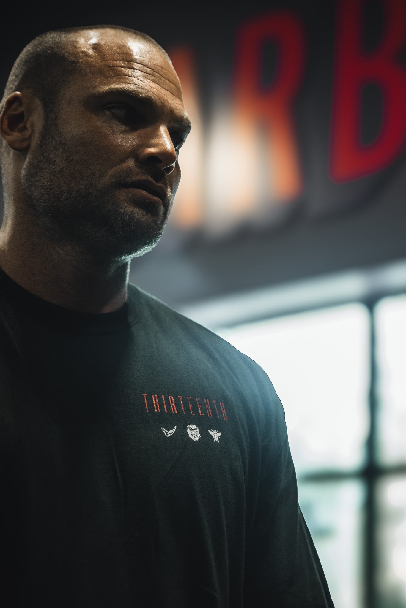 A man with a shaved head stands indoors before a blurred window, wearing the 13th Samson Premium Tee featuring THIRTEENTH and small graphics. The dim background shows red, out-of-focus letters.