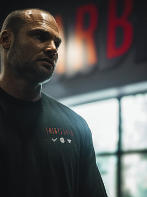 A man with a shaved head stands indoors before a blurred window, wearing the 13th Samson Premium Tee featuring THIRTEENTH and small graphics. The dim background shows red, out-of-focus letters.