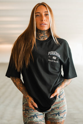 A woman with long brown hair and visible tattoos wears the Tango Charlie Apparel Persevere Premium Oversized Tee in black, made from heavyweight cotton. Indoors, she pairs it with patterned shorts and holds the hem of her shirt with both hands.