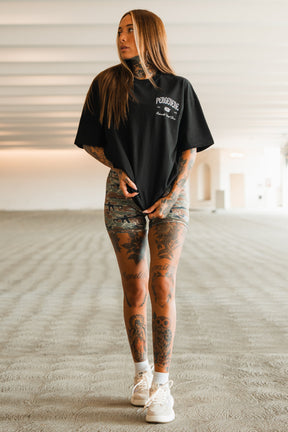 A woman with long hair and tattoos stands in a spacious, well-lit room. She wears the Persevere Premium Oversized Tee by Tango Charlie Apparel, paired with patterned shorts, white socks, and sneakers, gazing off to the side while holding her shirt.