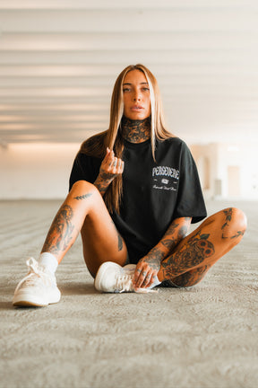 A tattooed woman with long hair sits in a relaxed pose, wearing the Tango Charlie Apparel Persevere Premium Oversized Tee and white sneakers, in a brightly lit, empty indoor space.