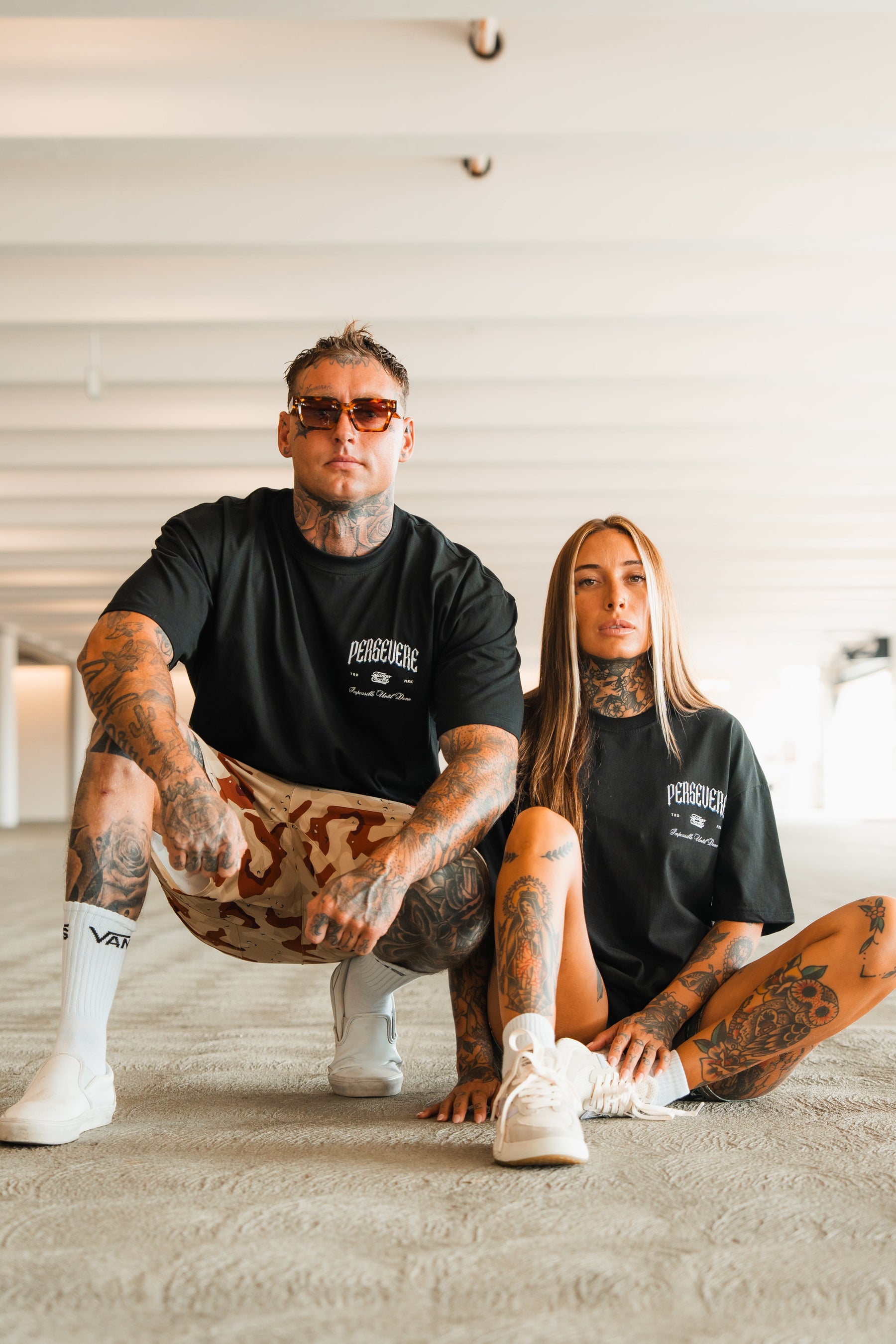 Two tattooed individuals confidently pose indoors wearing black Persevere Premium Oversized Tees by Tango Charlie Apparel and sneakers. One squats in sunglasses and shorts; the other sits with long hair and white shoes.