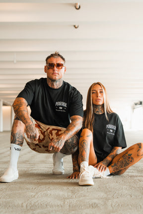 Two tattooed individuals confidently pose indoors wearing black Persevere Premium Oversized Tees by Tango Charlie Apparel and sneakers. One squats in sunglasses and shorts; the other sits with long hair and white shoes.