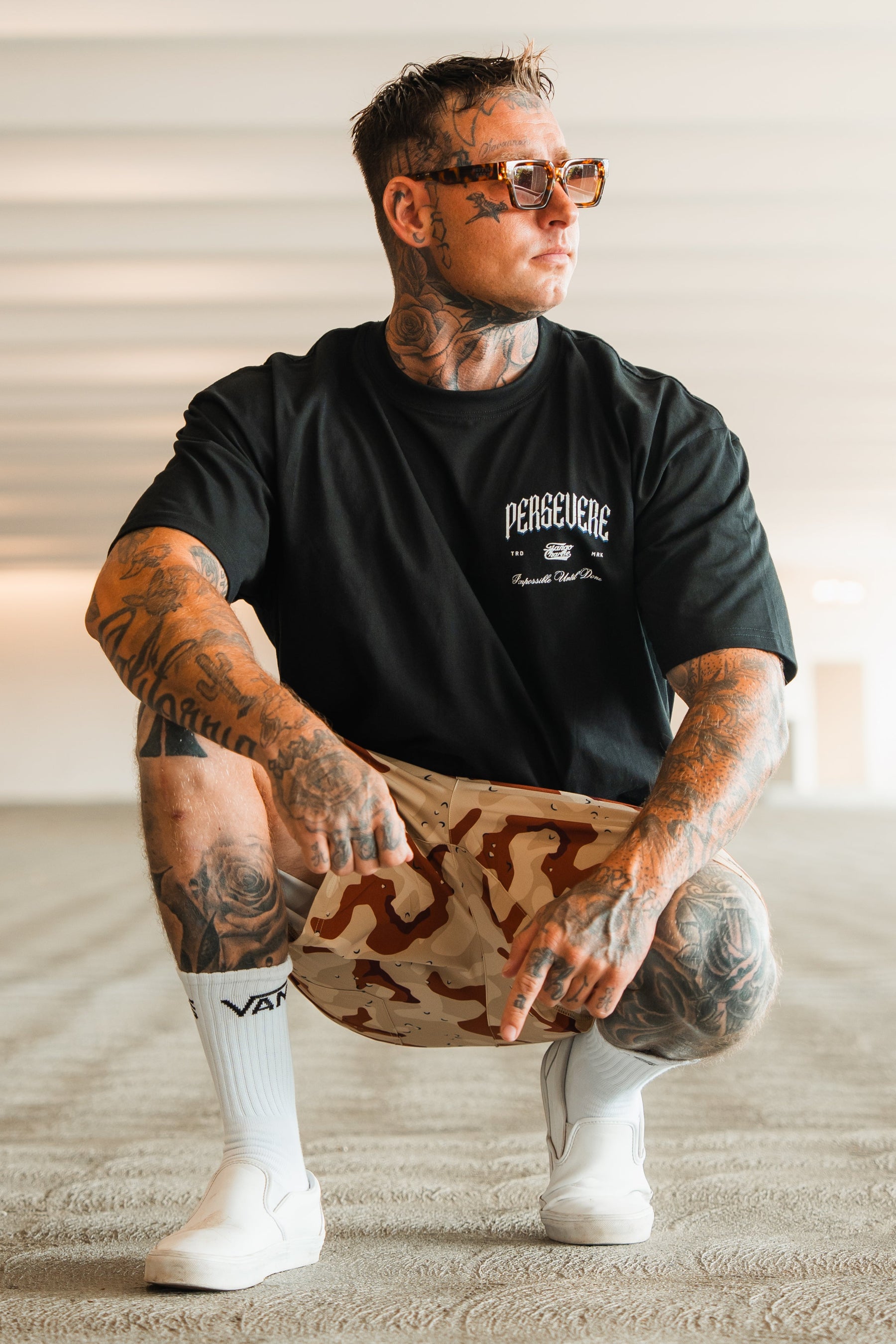 A tattooed man in sunglasses, crouching on a textured floor in a brightly lit modern space, wears the Tango Charlie Apparel Persevere Premium Oversized Tee with beige camo shorts and white slip-on shoes.
