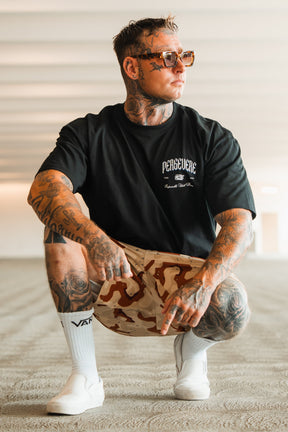 A tattooed man in sunglasses, crouching on a textured floor in a brightly lit modern space, wears the Tango Charlie Apparel Persevere Premium Oversized Tee with beige camo shorts and white slip-on shoes.