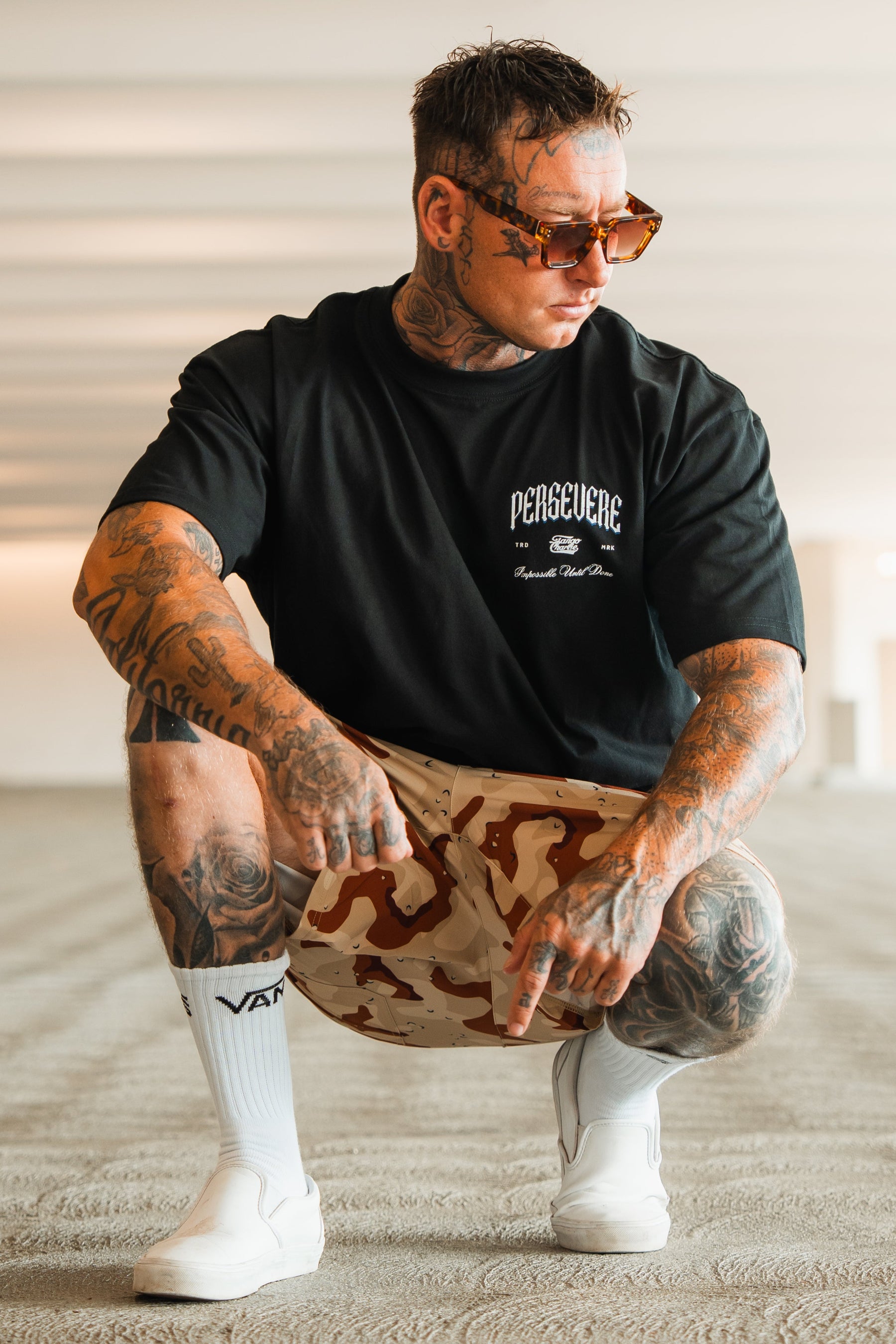 A tattooed man in sunglasses, camouflage shorts, white socks, and slip-on shoes squats indoors, pointing down and looking to the side while wearing the Tango Charlie Apparel Persevere Premium Oversized Tee.