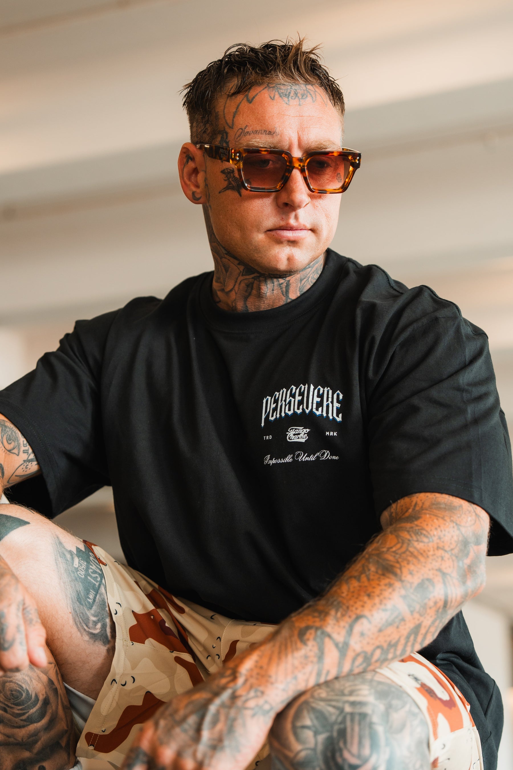 A tattooed man in sunglasses wears the Persevere Premium Oversized Tee by Tango Charlie Apparel, made from heavyweight cotton, and tan camo shorts. He sits indoors with a confident expression.