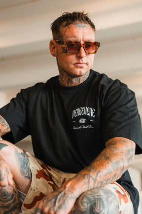 A tattooed man in sunglasses wears the Persevere Premium Oversized Tee by Tango Charlie Apparel, made from heavyweight cotton, and tan camo shorts. He sits indoors with a confident expression.