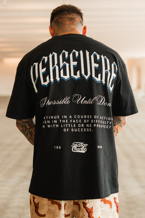 A tattooed man stands indoors in the Persevere Premium Oversized Tee by Tango Charlie Apparel—made from heavyweight cotton, it features bold “PERSEVERE,” “Possible Until Done,” and other motivational text. He pairs it with tan patterned shorts.