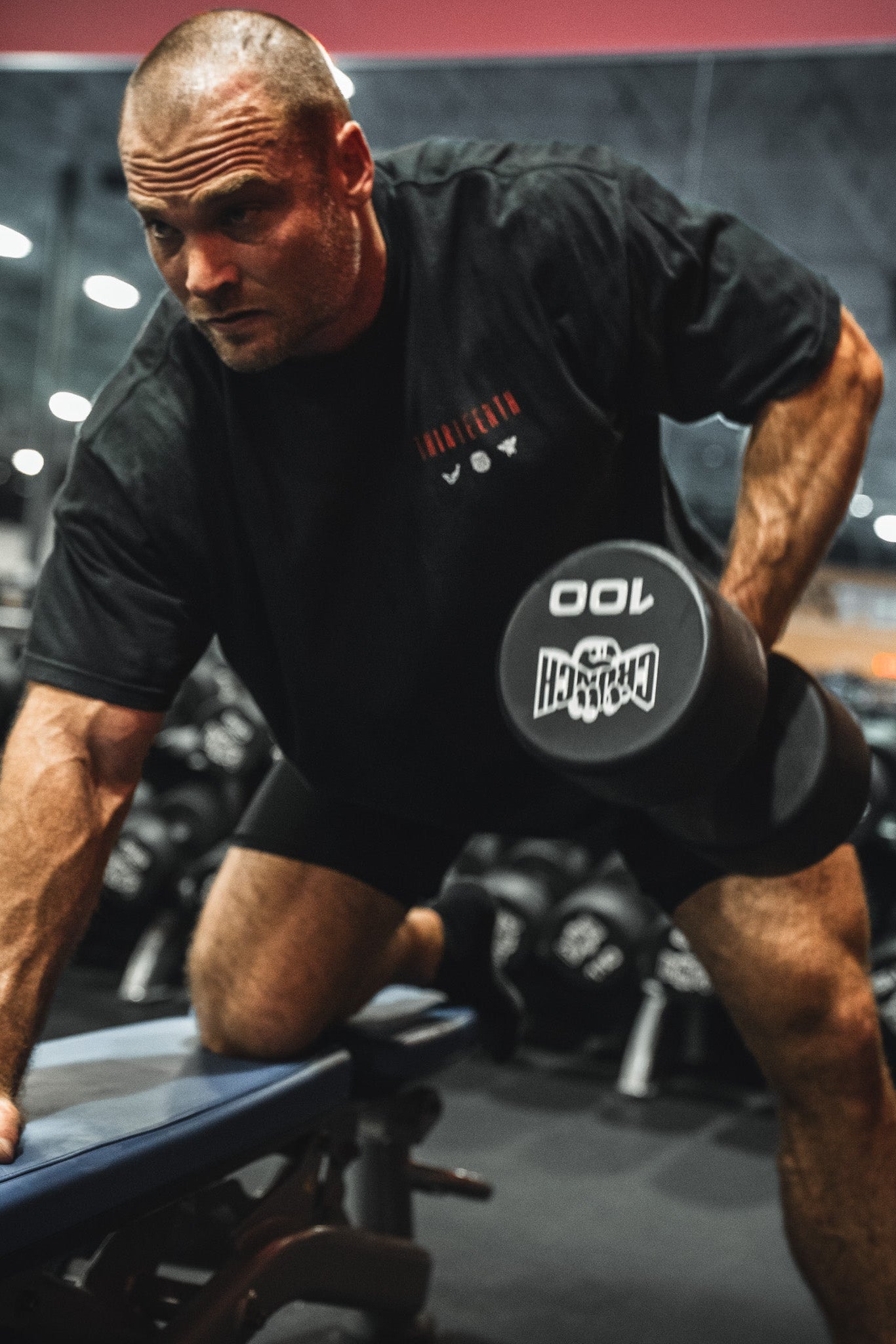A muscular man in a black 13th Samson Premium Tee and shorts performs a one-arm dumbbell row with a 100-pound weight on a gym bench, surrounded by mirrors and racks of dumbbells.