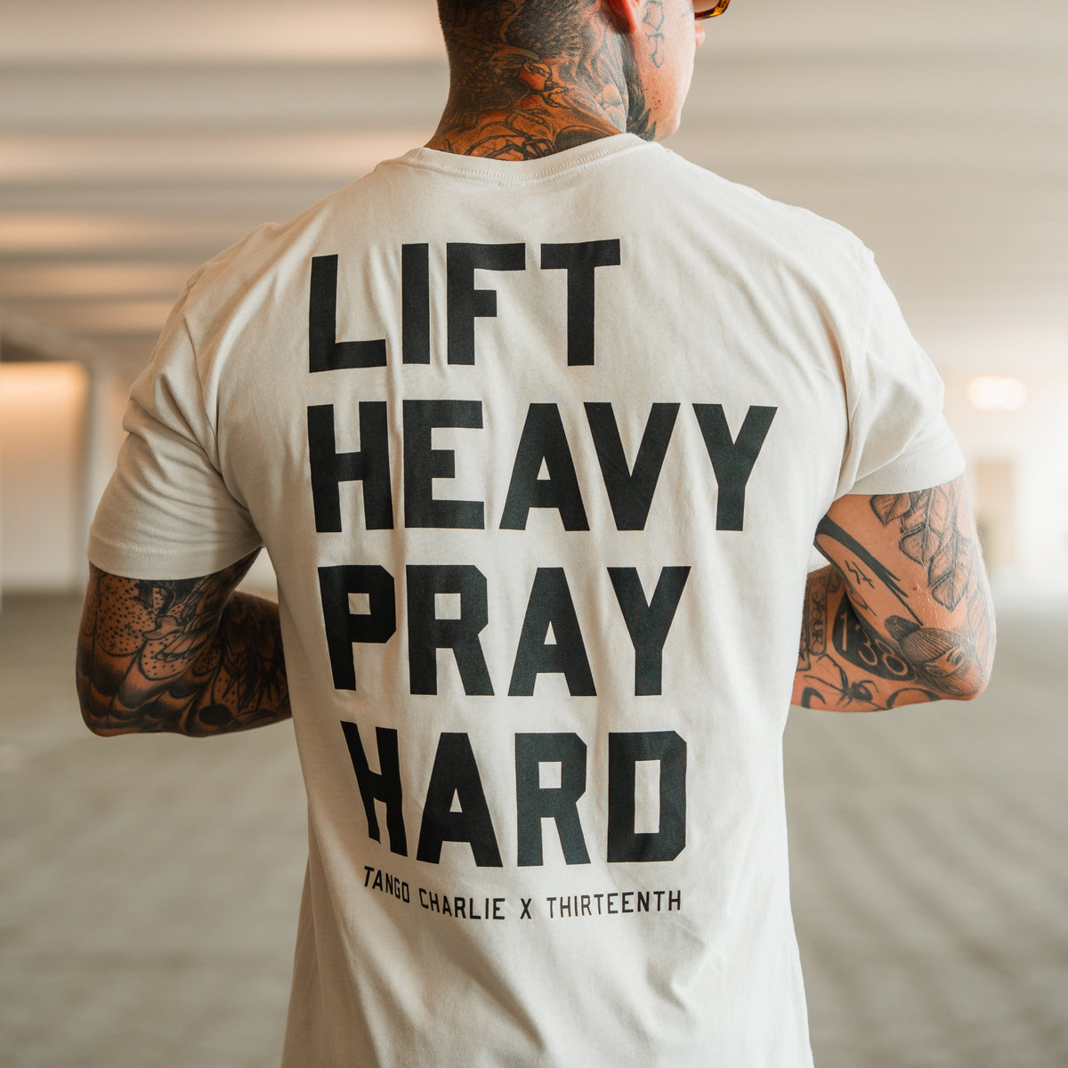 A tattooed person from behind wears a light tee with bold black LIFT HEAVY. PRAY HARD. text, symbolizing resilience. Below is TANGO CHARLIE APPAREL, denoting the brand and product: Lift Heavy. Pray Hard. - Tee by Tango Charlie Apparel.