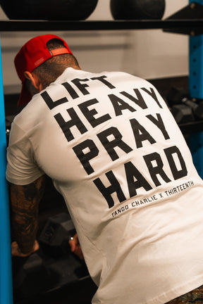 Wearing the Tango Charlie Apparel Lift Heavy. Pray Hard. tee and a red cap, someone leans over gym equipment, ready to lift, representing strength and faith.
