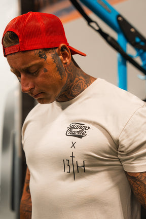 A tattooed man in a red backward cap looks down, wearing the white Lift Heavy. Pray Hard. - Tee by Tango Charlie Apparel, featuring bold black graphics. Blue and black shapes blur in the background.