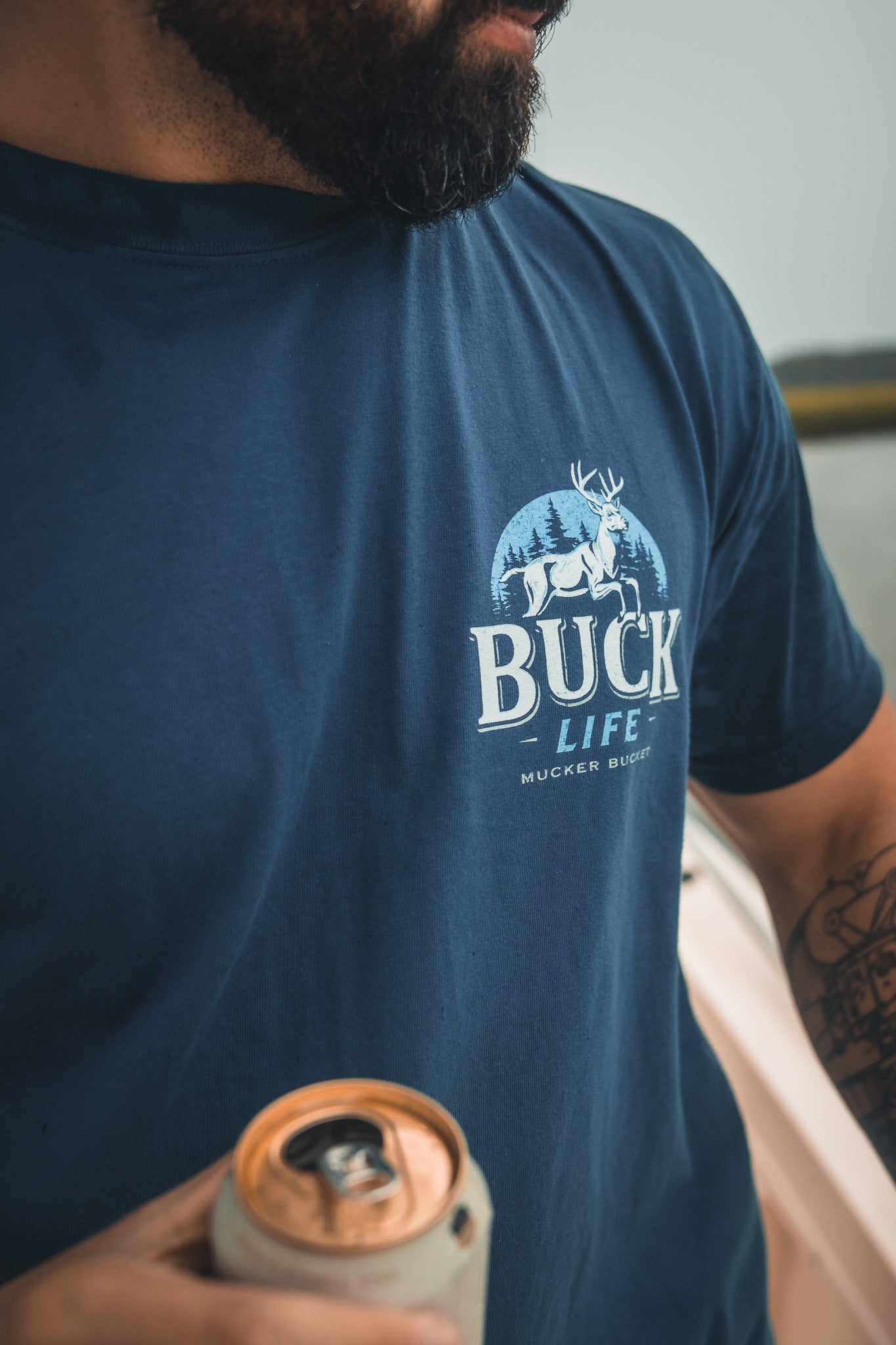 A bearded person holds a can in their tattooed hand, wearing a blue Mucker Bucket Buck Life Premium Tee with a white deer and trees graphic. Their face is partially out of frame.