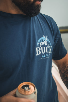 A bearded person holds a can in their tattooed hand, wearing a blue Mucker Bucket Buck Life Premium Tee with a white deer and trees graphic. Their face is partially out of frame.