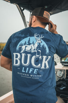 A man in a Mucker Bucket Buck Life Premium Tee, dyed blue and featuring a deer-in-forest graphic, stands on a boat adjusting his cap.
