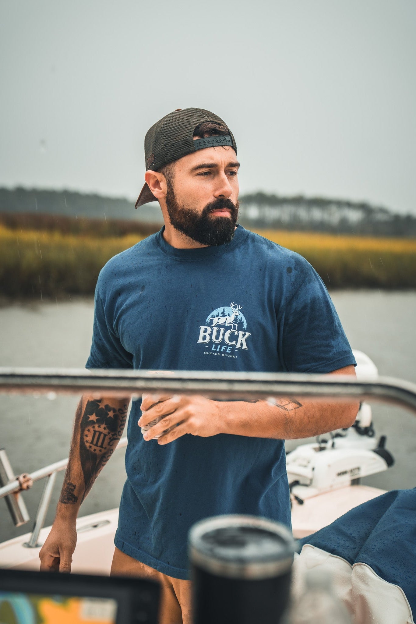 A bearded man in a blue Mucker Bucket Buck Life Premium Tee and backwards cap stands on a boat holding a drink, with marshland and water in the background.